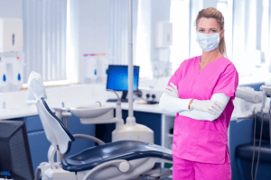 A woman in pink scrubs and white gloves standing next to a dentist 's chair.